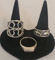 Three silver rings displayed on a black ring holder, showing different designs and surface wear.