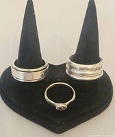 Three silver rings displayed on black ring holder, front and side profiles shown.
