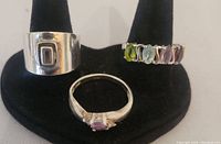 Three sterling silver rings displayed on a black ring holder, showing the wide band with rectangular design, the central pink stone ring, and the multi-colored gemstone band ring.
