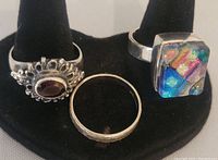 Photo showing the three rings displayed on a black holder, includes the red stone ring with ornate design, plain band, and the adjustable dichroic glass ring.