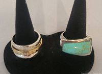 Two silver rings displayed on black ring holders, one with turquoise stone, one hammered band with gold accent