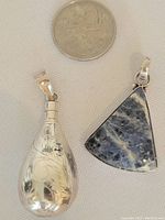 Two silver pendants side by side with coin for size reference; one is engraved with floral designs on silver teardrop shape, other is a triangular stone set pendant in blue and white colors.