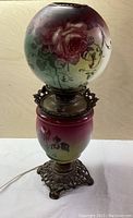Full view of antique Victorian lamp showing the painted glass globe with roses on top, and the painted glass body with matching rose design above cast brass base with ornate feet.