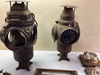Two cylindrical cast metal lanterns side by side on white surface against plywood background