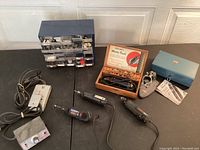 Photo shows the four Dremel rotary tools, one in a vintage wooden box with bits, plus clamping accessories in a blue box, arranged on a black surface.