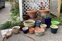 Overview of all planters including terracotta, ceramic, glazed, and decorative pots arranged on pavement beside white lattice screen.