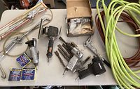 Photo showing collection of pneumatic tools on white surface including air drills, sander, grinder with sanding disc, several drill bits, cut-off wheels, and coiled hoses in yellow-green and brown.