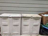 Two closed plastic yard storage containers side by side against a wall, showing front panel design and dirt.