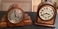Full view of two vintage wooden mantle clocks side by side on a black surface. The clock on left is wider with a wooden base and rounded top, the clock on right is taller with a more vertical rectangular wooden case and rounded top.