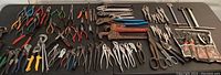 Wide shot of various hand tools arranged on a black surface, showing pliers, snips, cutters with colorful handles and several large pipe wrenches.