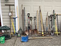 Photo showing a collection of garden tools lined up by garage door including shovels, rakes, post hole diggers, and other long-handled yard tools.