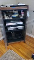 Wooden media cabinet with glass doors holding three electronic components on shelves plus remotes and cables on top.