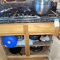 Shows stove top with Calphalon non-stick pan, large stainless steel mixing bowl, and cabinet below with cast iron pans, pots, and kitchen items including blue silicone funnel.
