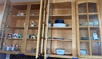 Wooden cabinetry with glass doors open exposing various Corning Ware cookware, mugs, plates, and glassware inside