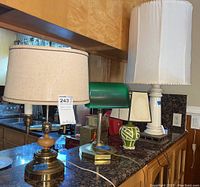 Four lamps on dark granite countertop including brass and wood lamp, green banker's lamp, green and white ceramic lamp, and tall cream ceramic lamp with fabric shade.