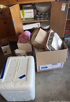 Full view of lot with cardboard boxes, styrofoam cooler, and wooden cabinet containing kitchenware items.