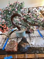 Cherry blossom tree in a light green ceramic pot, natural decorative rocks, and carved metal box on a glass table with lace tablecloth.