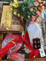 Photo of several decor items on a table including boxed black carved figure, red hanging wall ornaments, and bunches of artificial flowers