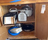 Photo showing assortment of kitchenware items on wooden shelf including rice cookers, kitchen scale, blue baking dish, metal pan, and white plastic item