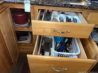 Kitchen open drawers showing wooden cabinetry with cutlery and utensils in white trays and red ceramic pot.