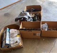 Five cardboard boxes containing various outdoor plumbing parts, extension cords, sprinkler fittings, and caster wheels on cement floor.
