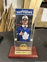 Full front view showing Auston Matthews photo card inside clear acrylic case with bronze medallion below and wooden base with Toronto Maple Leafs plaque.