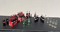 Wide view of grouped Coca Cola bottles on table showing multiple filled bottles in cardboard carriers, empty green bottles, and clear empty bottles