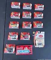 Photo showing all 13 Coca Cola branded collectible vehicles in original packaging arranged on black surface, including sports cars, vans, SUVs, and transport trucks.