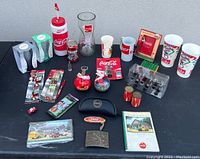 Overview of Coca Cola collectible items arranged on a black surface including cups, ornaments, collectible pencils, and tin items.