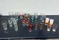 Wide shot showing all 16 glasses arranged on a black table outdoors revealing different branded and styled vintage drinking glasses.