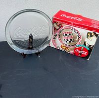 Glass serving plate with embossed Coca Cola logo on a stand alongside the boxed 12 piece dinnerware set