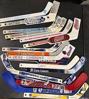 Photo showing a collection of 12 mini hockey sticks with various team and sponsor logos, blades in blue, red, white, and wooden finishes.
