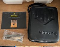 Black carrying case with Moozica logo, instruction booklet for kalimba, and bag with metal parts including tone hammer.