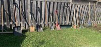 Outdoor tools lined up against wooden fence including shovels, rakes, brooms