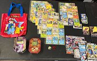 Full view of the Pokémon cards spread out on table, two Pokémon-themed metal tin storage cases, and red Pokémon bag with Ash and Pikachu graphic.
