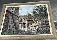 Framed oil painting of a cobblestone courtyard and stone houses with leafy trees and hills in the background