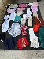 Photo showing a variety of baby clothing laid out on black surface including onzies, dresses, shirts, hats, and pants in multiple colors and patterns.