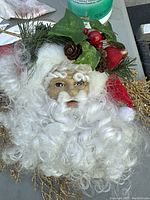 Santa Claus face wreath with white curly hair, foliage, pine cones, and holiday decor