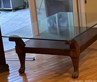 Large wood coffee table with thick glass top and carved legs, showing detail of woodwork and glass.