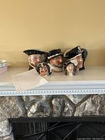 Five Royal Doulton character mugs in various sizes and detailed painted faces on a white mantel.
