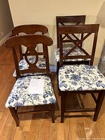 Four wooden dining chairs displayed, two tall with cross-back design and two shorter with curved slat back, all with blue floral fabric seats.
