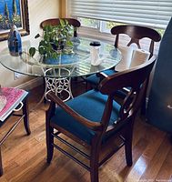 Round glass dining table with white metal base and four wooden chairs with blue cushions, two chairs with arms and two without; setup in a sunlit room with hardwood floor.