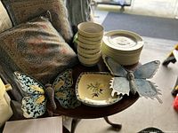 Image showing stacks of Royal Doulton fine china cups and plates alongside a floral serving dish, four patterned decorative pillows, and two large metal butterfly decorations on a wooden surface.