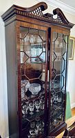 Front view of dark wood china cabinet with glass doors and intricate geometric wooden pattern, showing glass shelves inside with crystal glassware.