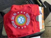 Red pet cape with Superdog graphic and text, new with packaging visible.