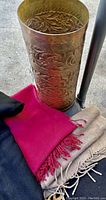 Multiple scarves in various colors next to a tall brass tone holder with embossed floral or abstract design.