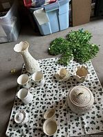 Overview of the lot showing ceramic vase, mugs, bowls, containers, tea cup and saucer, coffee cup and shamrock fabric towels, and shamrock greenery wall hanging lying on a shamrock-themed towel on the floor.