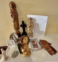 Photo showing various carved wood religious figurines including Madonna and Child statues, a black cross statue, wooden hand carving with prayer, and small framed items.