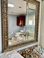 Large rectangular mirror with ornately carved gold-tone frame hung on a bathroom wall above a sink.