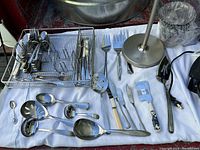 Metal kitchen utensils organized in a metal rack including forks, spoons, knives, and serving spoons next to a cutting board rack and lamp base.